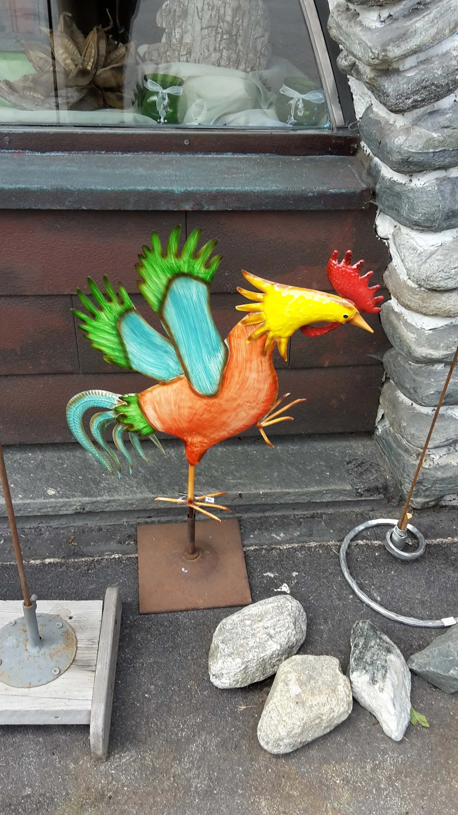 Farbig gefasster geschmiedeter Hahn auf einem Ständer vor dem Schaufenster des Ladens